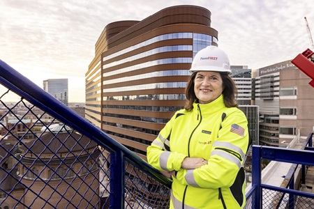 Kathy Gallagher, MSN, clinical liaison for the Pavilion, stands with arms folded wearing a yellow vest and hard hat on the roof of HUP with the Pavilion in the background.